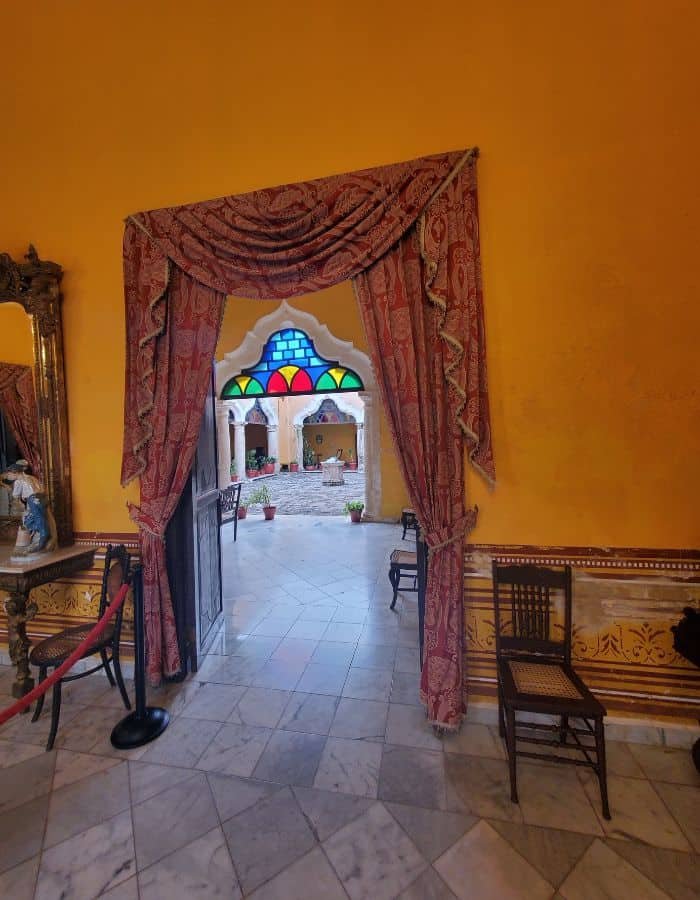 Interior of a historic museum in Campeche with gold-colored walls and a doorway framed by elegant red drapes. Through the doorway, a room with colorful stained glass windows, tiled floor, and colonial-style furnishings is visible.