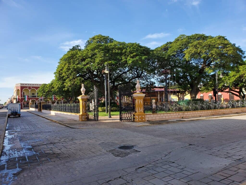 Charming corner park in Campeche, Mexico, enclosed by a black wrought-iron fence with stone pillars. Lush green trees provide dense shade over benches and walkways, set against a backdrop of colonial buildings and a clear blue sky.