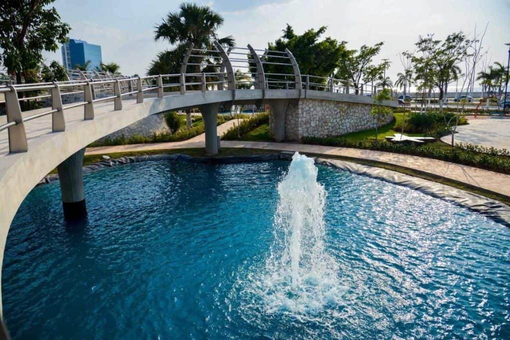 Modern park in Campeche featuring a circular fountain pool with a central water jet and an arched pedestrian bridge above. Palm trees and landscaped paths surround the serene blue water under a partly sunny sky.