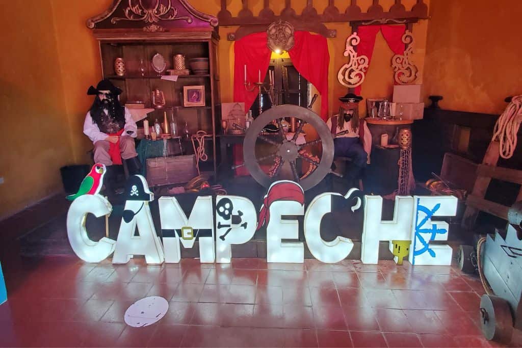 Indoor pirate-themed display with large, decorated “Campeche” letters in front of costumed mannequins, props like a ship’s wheel, and nautical decor. The setup includes red curtains, pirate flags, and various historical artifacts, creating a playful, museum-like atmosphere.