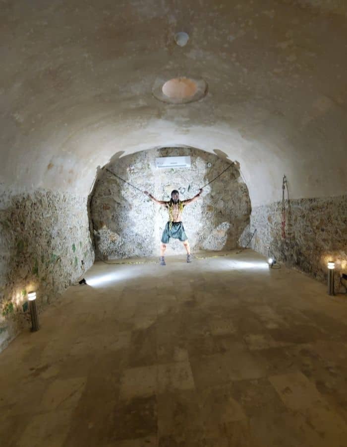 Dimly lit stone chamber with a life-sized mannequin of a chained prisoner posed against the far wall. The arched ceiling and rough stone walls suggest a historic dungeon or jail, with small lights adding dramatic effect.
