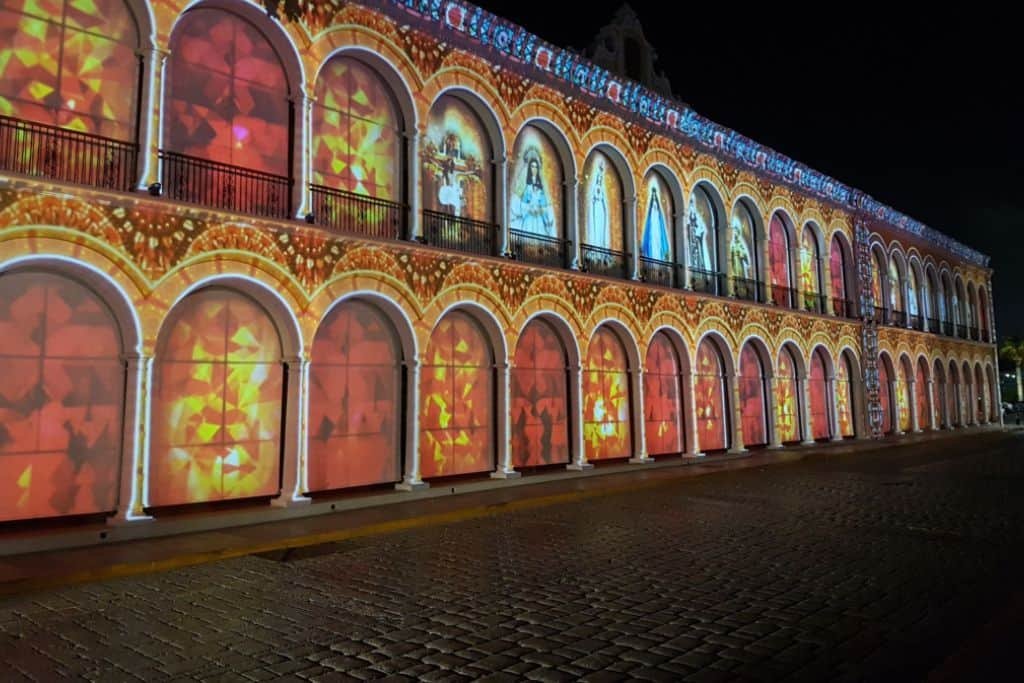 Colonial-style building in Campeche illuminated at night with vibrant light projections filling its arched windows and walls. The colorful visuals create a festive, stained-glass effect across the entire façade, likely part of a public light show or cultural event.
