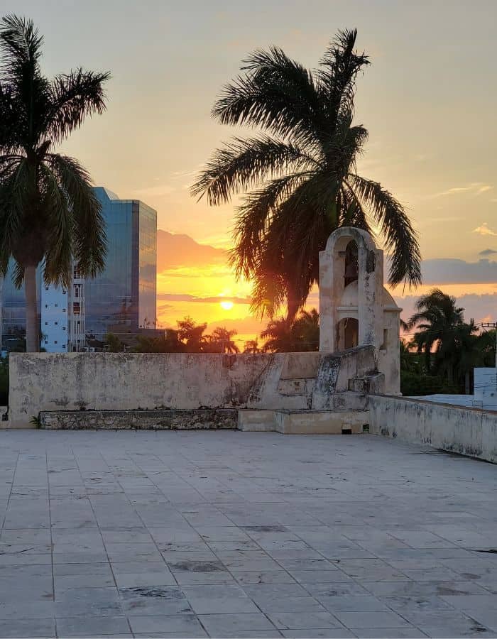 Sunset view from a historic rooftop in Campeche, featuring palm trees silhouetted against the golden sky. A weathered stone bell tower stands on the right, with modern buildings peeking through the tropical landscape in the background.