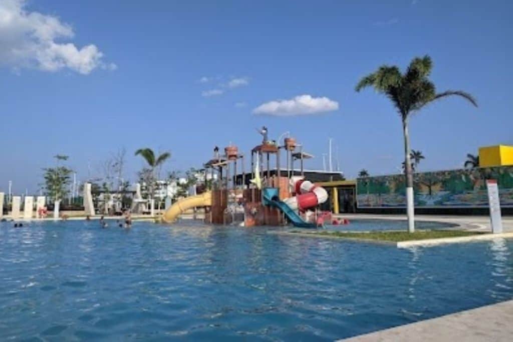 Outdoor water park in Campeche featuring a large pool and colorful pirate-themed play structure with multiple slides. Palm trees and a bright blue sky create a tropical, family-friendly atmosphere.