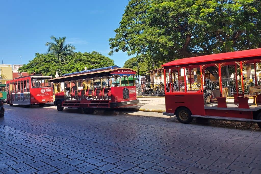 Three bright red open-air trolleys lined up along a cobblestone street in a shaded area of Campeche. Trees, park benches, and colonial-style buildings are visible in the background under a sunny blue sky.