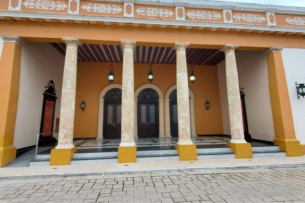 Exterior of a historic theater in Campeche, Mexico, featuring a covered portico supported by six stone columns. The façade is painted in warm tones of orange and cream, with decorative trim and arched doorways set above a checkered tile floor.