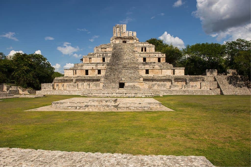 The grand stepped pyramid of Edzná, an ancient Mayan archaeological site in Campeche, Mexico, stands under a partly cloudy sky. The well-preserved limestone structure rises in multiple tiers with a central staircase, surrounded by grassy plazas and jungle trees.