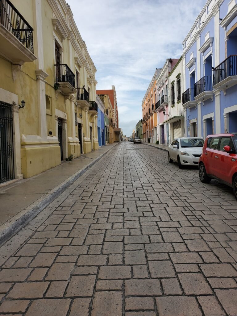 Quiet cobblestone street lined with colorful colonial-style buildings in shades of yellow, blue, and orange. A few parked cars sit along the sides, and the street stretches into the distance under a partly cloudy sky.