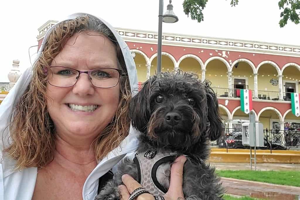 A smiling woman in a light hoodie poses with a small black dog in front of a colorful colonial-style building with arches and Mexican flags. The setting appears to be a plaza in Campeche, Mexico, with festive decorations and a relaxed, rainy-day vibe.