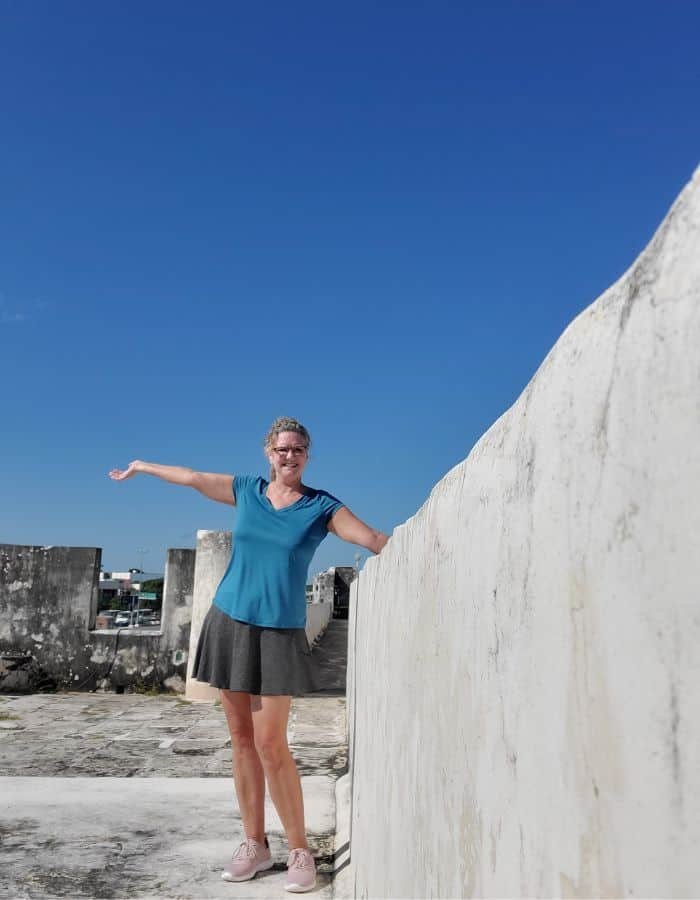 A woman in a teal top and gray skirt stands with one arm outstretched on a historic stone wall under a vivid blue sky. The setting appears to be a fortified area in Campeche, Mexico, with old stone structures visible in the background.