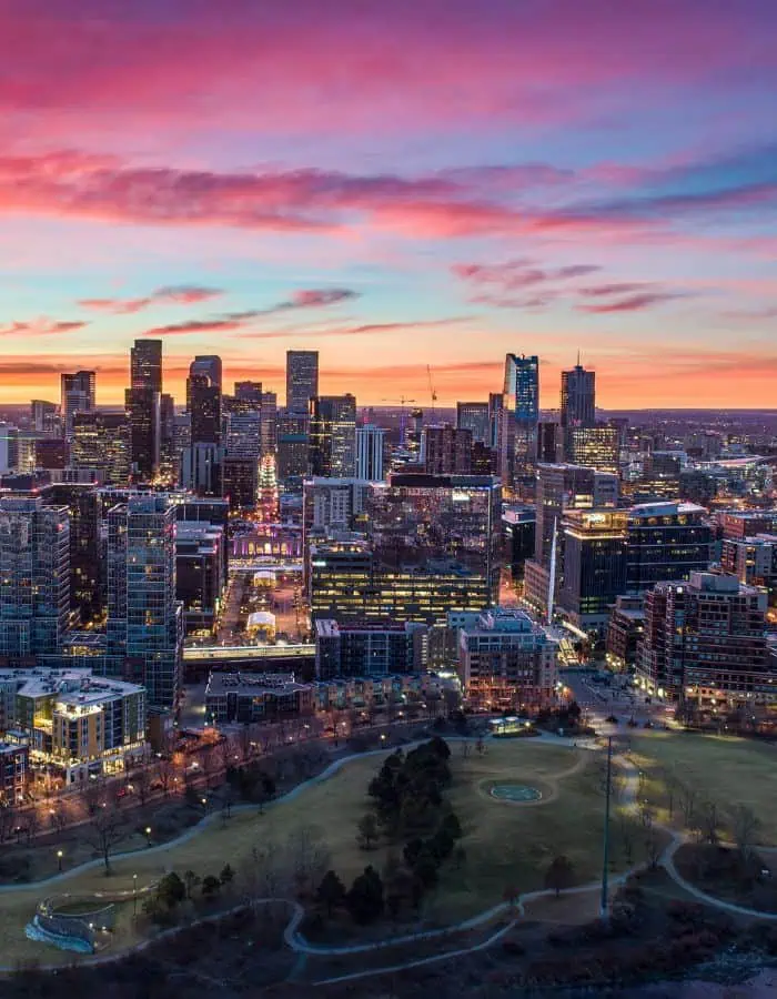 A vibrant aerial view of downtown Denver at sunset, with a dramatic sky streaked in pink, purple, and orange hues. The city’s skyline is lit with glowing windows and streetlights, while a large park with winding paths and clusters of trees sits in the foreground.