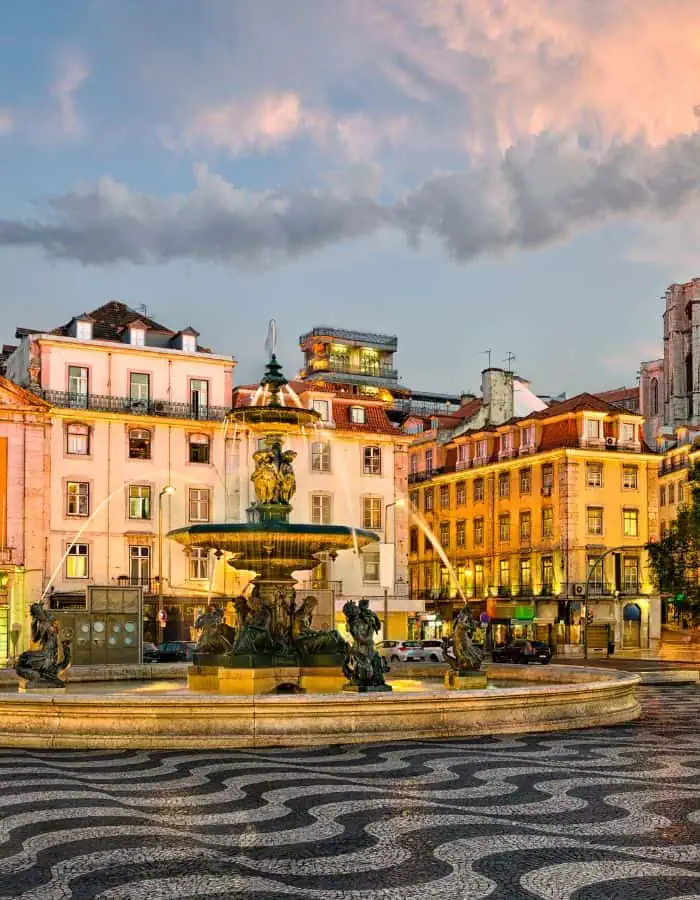 A beautifully lit fountain in Lisbon’s Rossio Square features ornate sculptures and arched streams of water, set against a backdrop of pastel-colored buildings with red-tiled roofs. The patterned black-and-white stone pavement adds charm to the scene as the sky glows with soft evening hues.