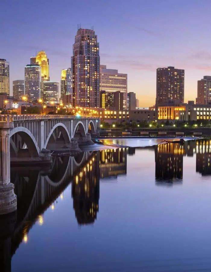 A twilight view of downtown Minneapolis, Minnesota, featuring a stone arch bridge lit with warm lights stretching across the calm Mississippi River. The city skyline glows against a dusky sky, with the buildings and bridge beautifully reflected in the water below.