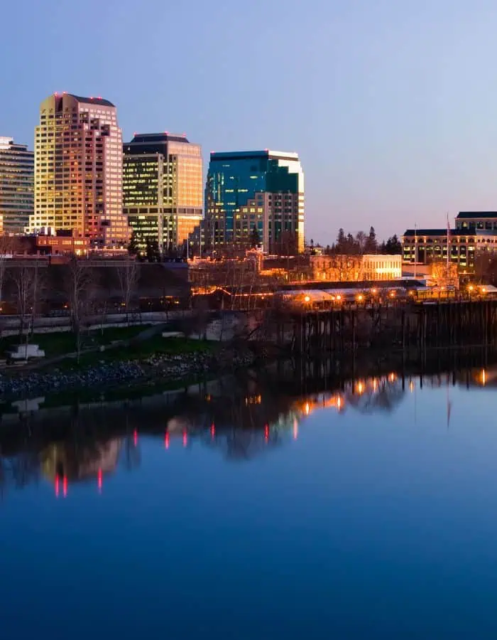 A tranquil dusk view of downtown Sacramento, California, with office buildings softly lit against a clear twilight sky. The city lights reflect gently on the smooth surface of the river, while bare trees and a wooden pier line the riverbank.