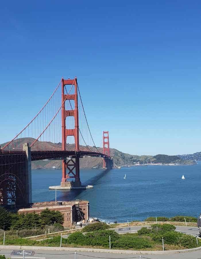 The iconic Golden Gate Bridge stretches across the blue waters of the San Francisco Bay under a clear sky. The rust-red towers rise prominently, with sailboats dotting the water below and rolling hills in the background.