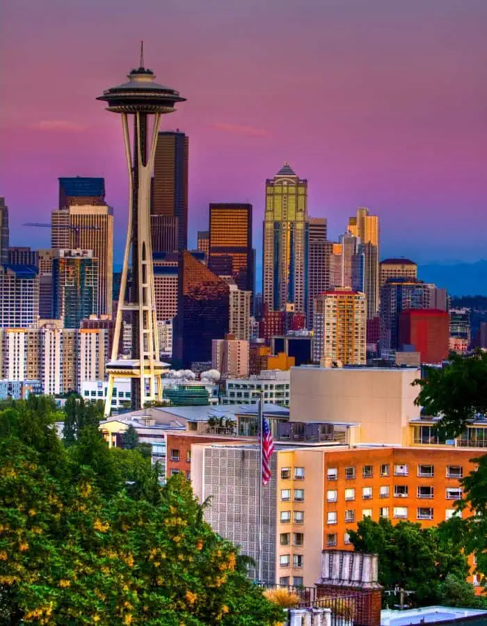 A vibrant view of downtown Seattle at sunset, with the iconic Space Needle prominently in the foreground. The city’s skyscrapers glow in warm evening light under a colorful sky blending shades of purple, pink, and orange, framed by lush green trees below.