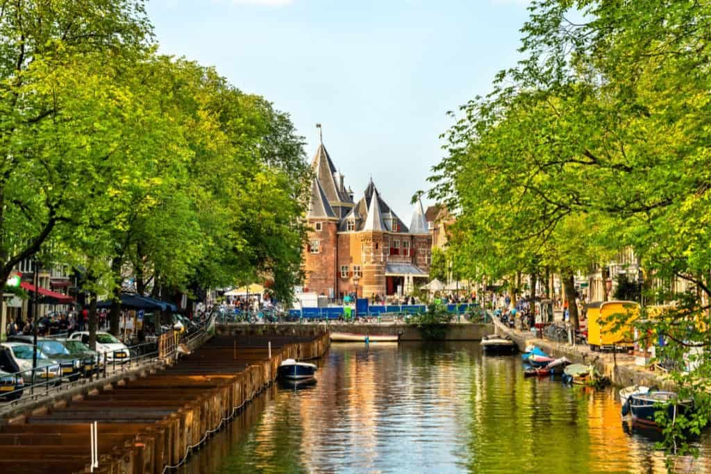 A charming canal in Amsterdam is lined with trees, parked boats, and quaint buildings, leading to the historic Waag building with its pointed towers. The water reflects the greenery and warm tones of the city, creating a peaceful and picturesque scene.