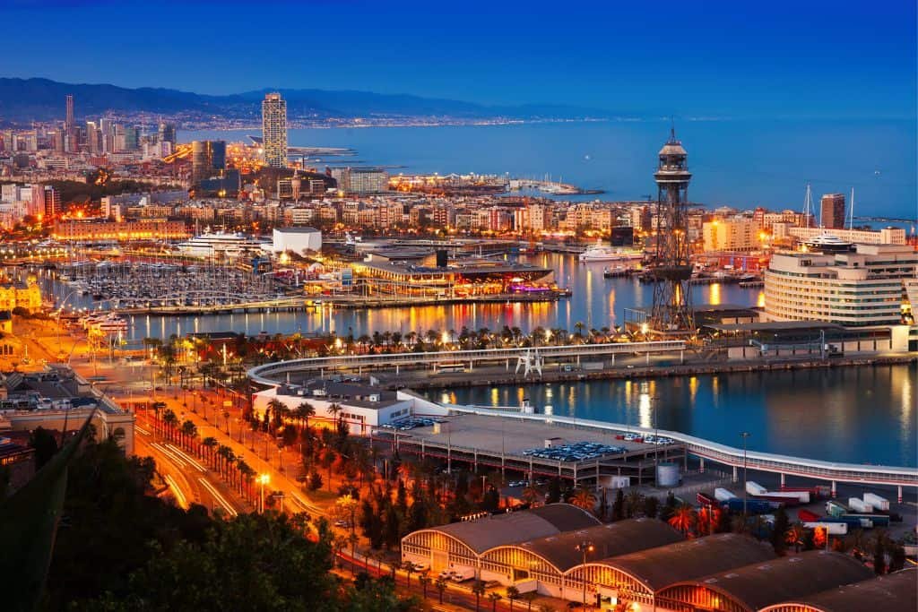 A stunning evening view of Barcelona’s waterfront, with city lights glowing along the harbor and modern buildings stretching into the distance. The Mediterranean Sea reflects the deep blue sky, and the city’s mix of historic charm and contemporary architecture is on full display.