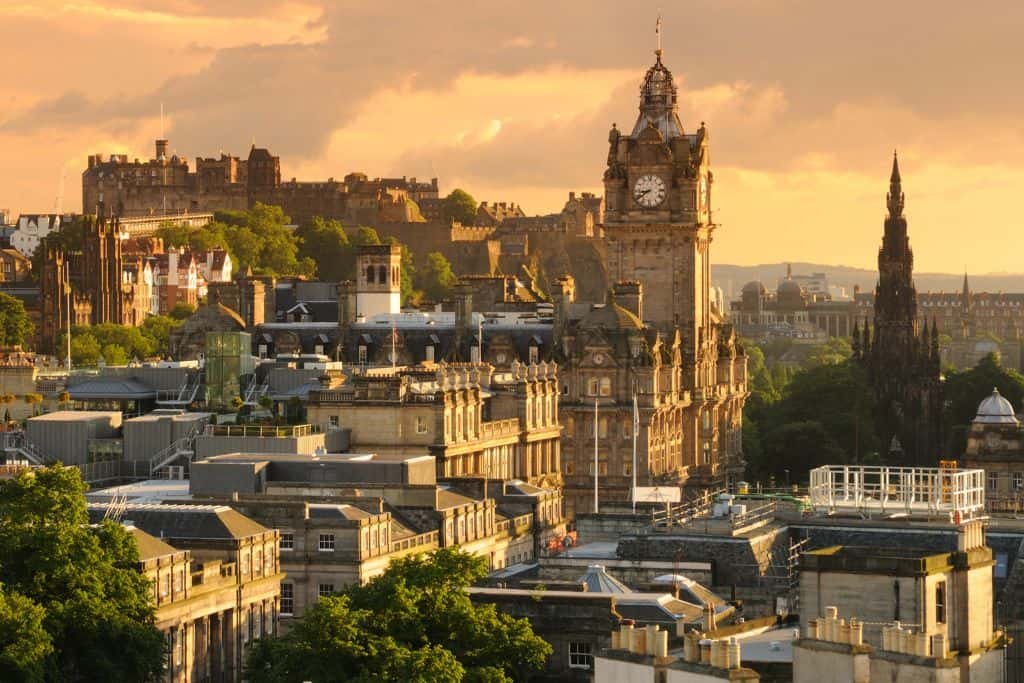 A golden sunset bathes the historic cityscape of Edinburgh, Scotland, highlighting the iconic Balmoral Clock Tower and the gothic spire of the Scott Monument. In the distance, Edinburgh Castle sits atop a rocky hill, overlooking the rooftops and winding streets below.