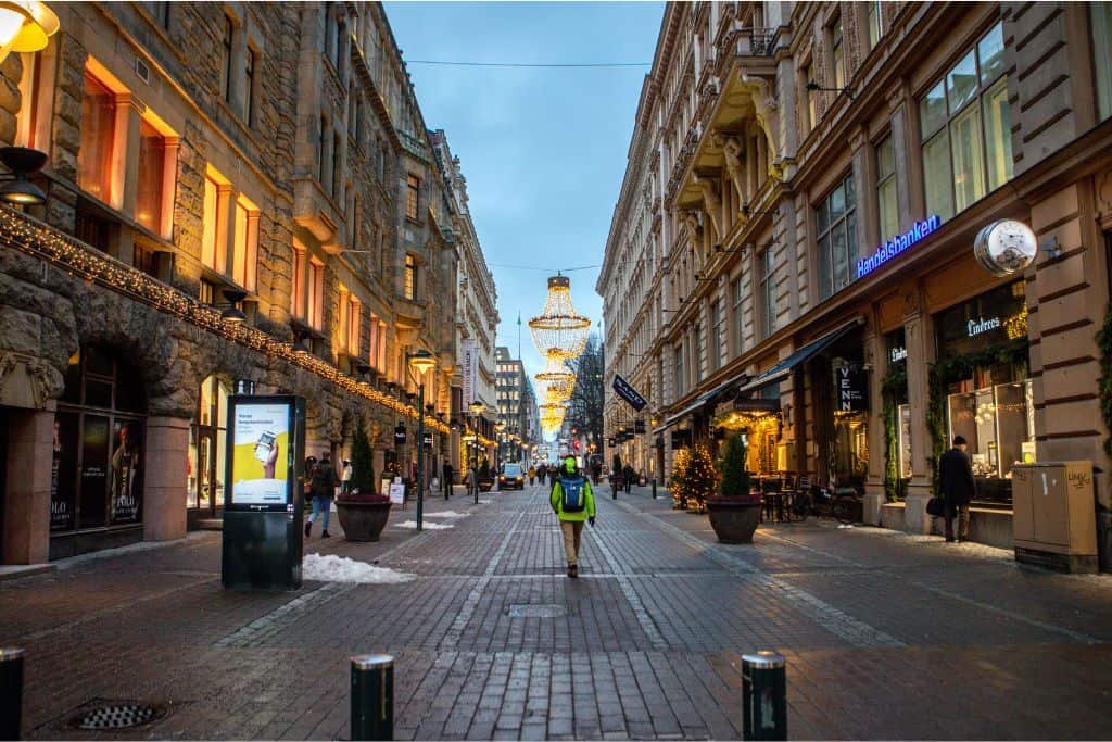A pedestrian street in Helsinki glows with warm lights from historic buildings and modern shops as evening sets in. A few people stroll along the cobblestone path beneath festive hanging lights, creating a cozy and inviting urban scene.