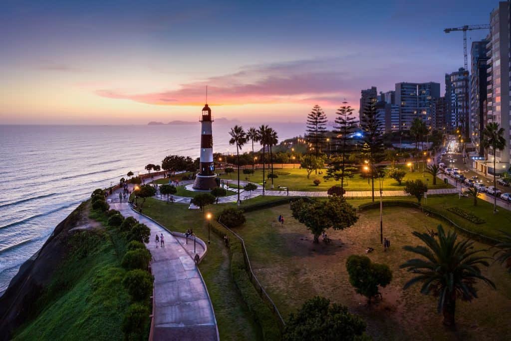 A scenic sunset view in Lima, Peru, features a black-and-white striped lighthouse perched on a coastal cliff, surrounded by a lush green park. The Pacific Ocean stretches out to the horizon, while modern high-rise buildings line the edge of the city in the background.