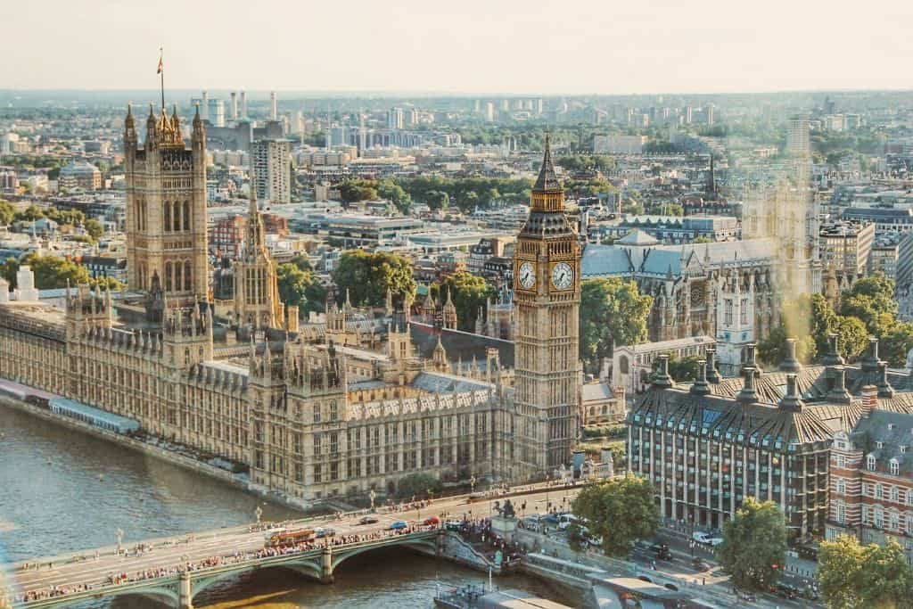 An aerial view of London highlights the majestic Houses of Parliament and the iconic Big Ben clock tower beside the River Thames. The Westminster Bridge stretches across the river, bustling with red double-decker buses and traffic, with the city's dense urban landscape extending into the distance.