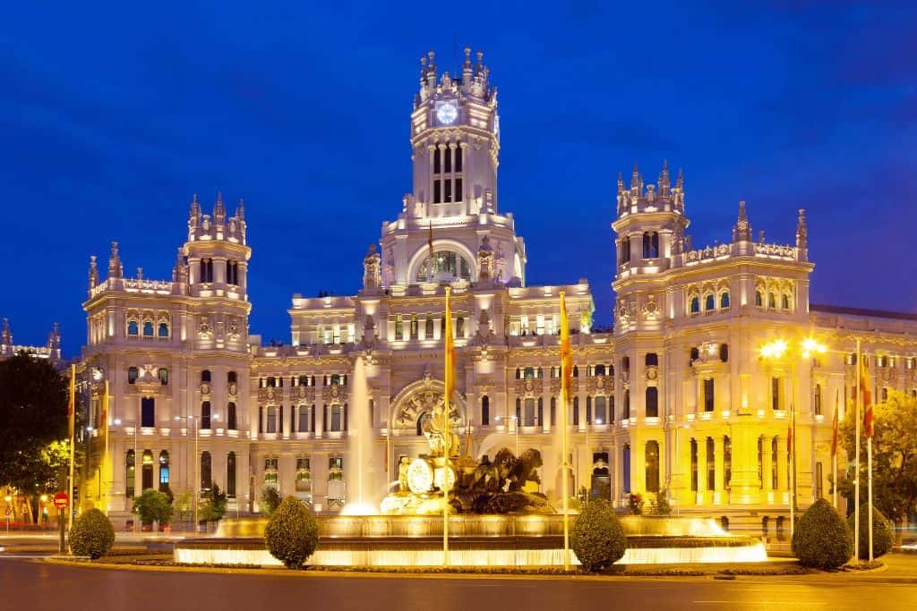 The Palacio de Cibeles in Madrid glows brilliantly at night, illuminated against a deep blue sky. The grand, ornate building is adorned with multiple towers and arched windows, while the iconic Cibeles Fountain stands prominently in front of it.