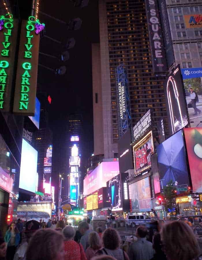 A bustling crowd walks through Times Square in New York City at night, surrounded by glowing billboards, neon lights, and advertisements—including a prominent Olive Garden sign. The scene captures the vibrant energy and bright chaos of the city after dark.