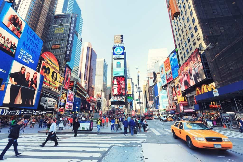 A bustling daytime view of Times Square in New York City, filled with people crossing the street and surrounded by colorful billboards and advertisements. A signature yellow taxi drives through the intersection, capturing the city’s fast-paced energy.