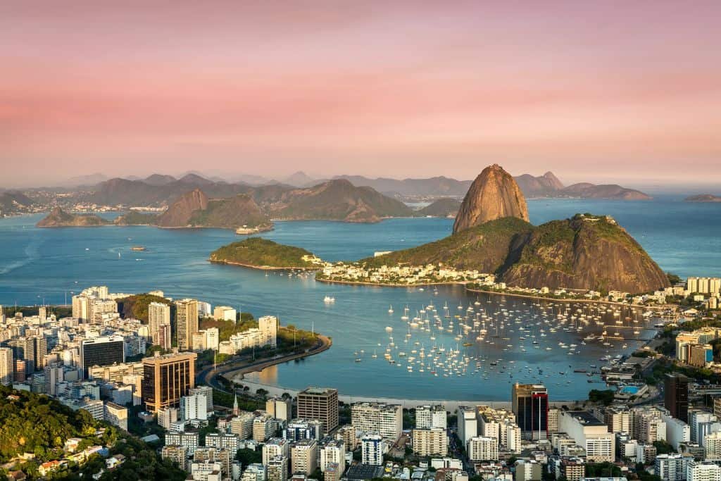 A breathtaking view of Rio de Janeiro at sunset, showcasing the iconic Sugarloaf Mountain rising above Guanabara Bay. The city’s buildings and marina are nestled along the coastline, with dozens of sailboats dotting the tranquil water under a soft pink sky.