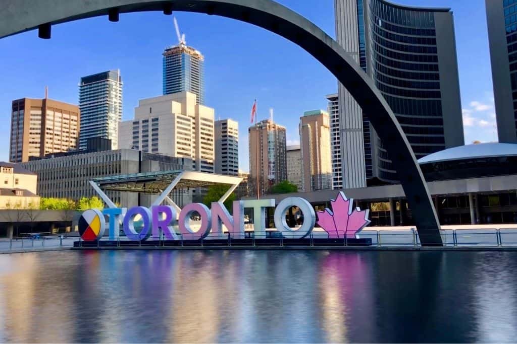 A colorful “TORONTO” sign sits in front of a reflecting pool at Nathan Phillips Square, with the modern towers of downtown Toronto rising in the background. A curved concrete arch spans the scene under a clear blue sky, capturing a bright and lively urban atmosphere.