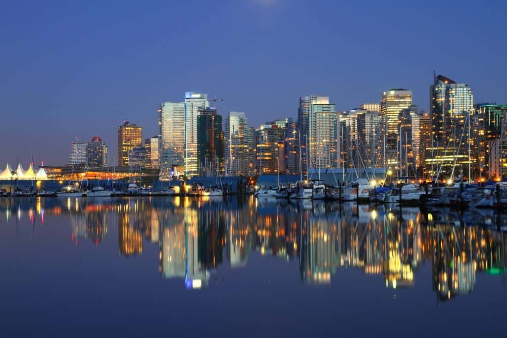 A dazzling view of Vancouver’s skyline at dusk, with glassy high-rises and waterfront buildings reflecting perfectly in the calm harbor. The lights from the city and anchored boats add a warm glow under the deepening blue sky.