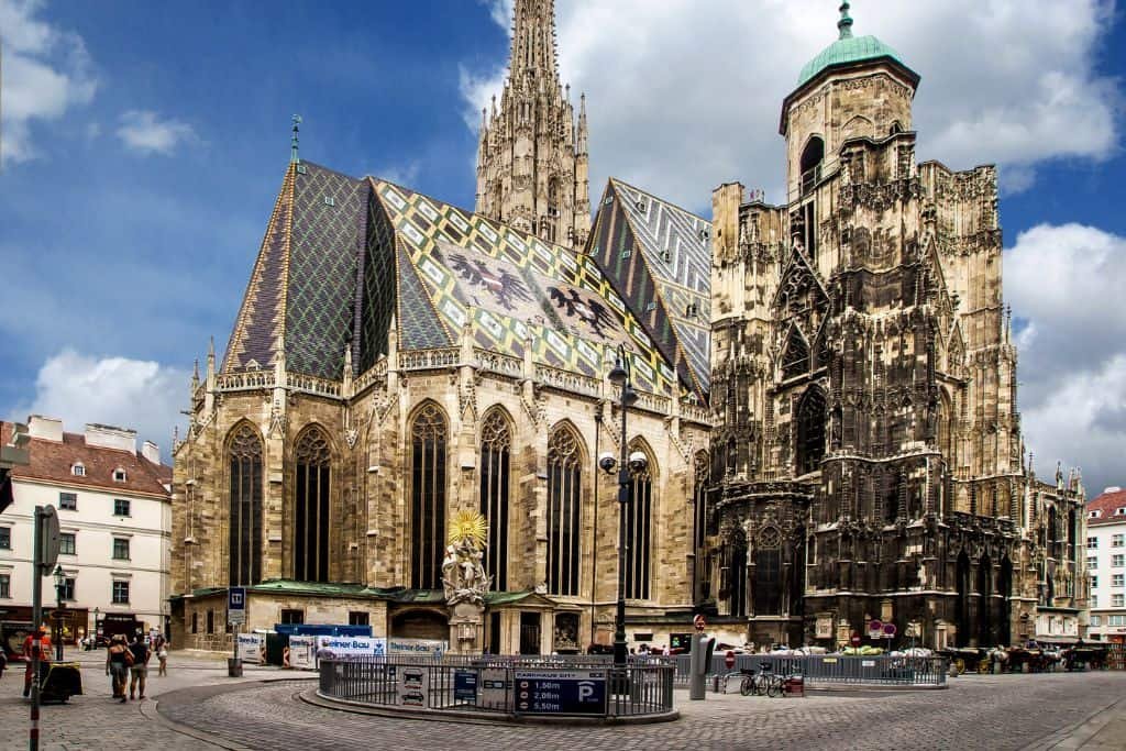 St. Stephen’s Cathedral in Vienna stands tall with its richly detailed Gothic architecture and colorful patterned tile roof. The cathedral's spire reaches high into the sky, surrounded by a quaint plaza with pedestrians and historic buildings.