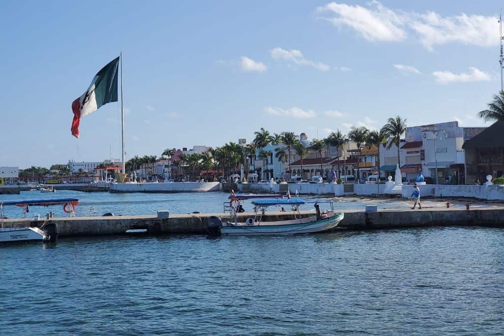 Waterfront scene in Cozumel featuring small boats docked along a pier, colorful buildings lining the shore, and a large Mexican flag waving prominently in the breeze under a sunny sky.
