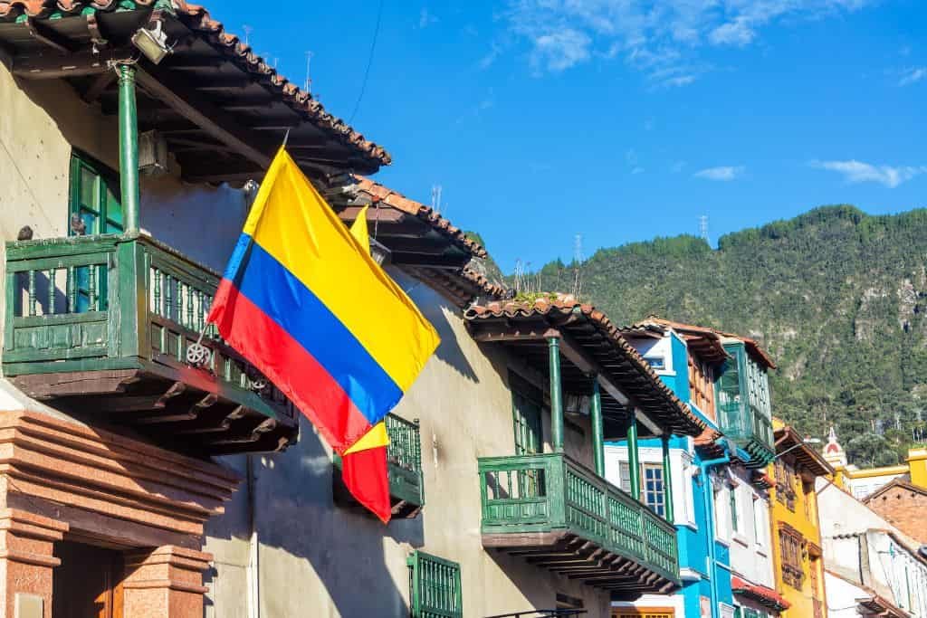 This image shows a Colombian flag hanging from a wooden balcony on a colorful colonial-style building. Bright blue skies and green hills provide a scenic backdrop to the vibrant street scene.