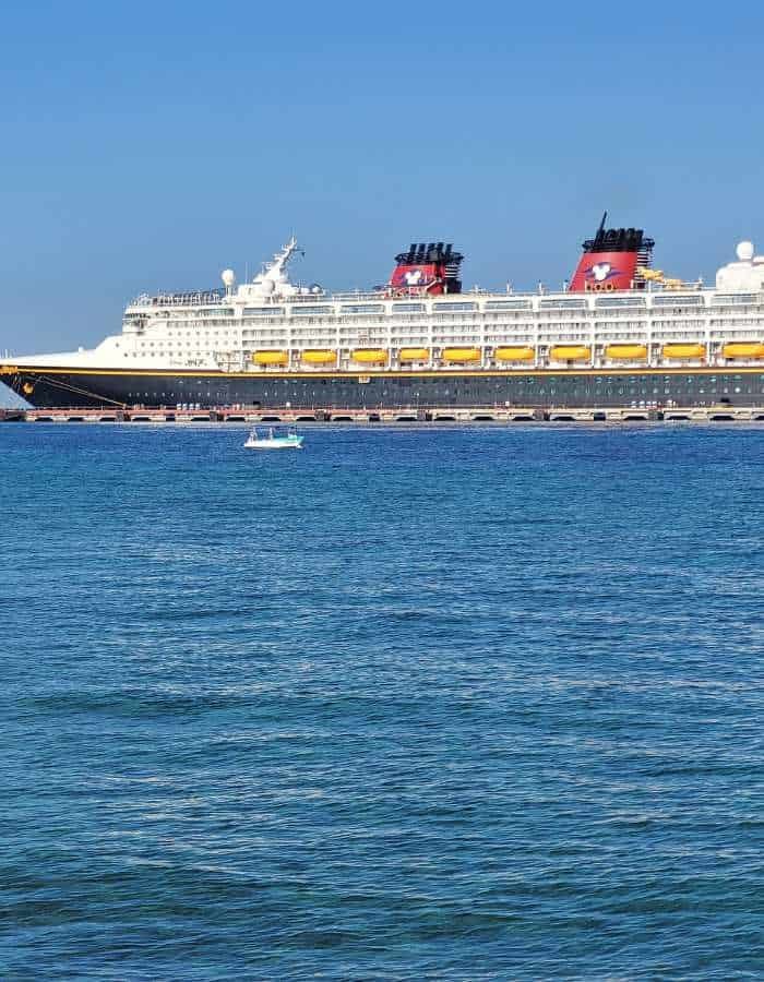 Large Disney cruise ship docked in Cozumel, featuring signature red and black funnels with Mickey Mouse logos, viewed from the calm blue waters of the Caribbean Sea.