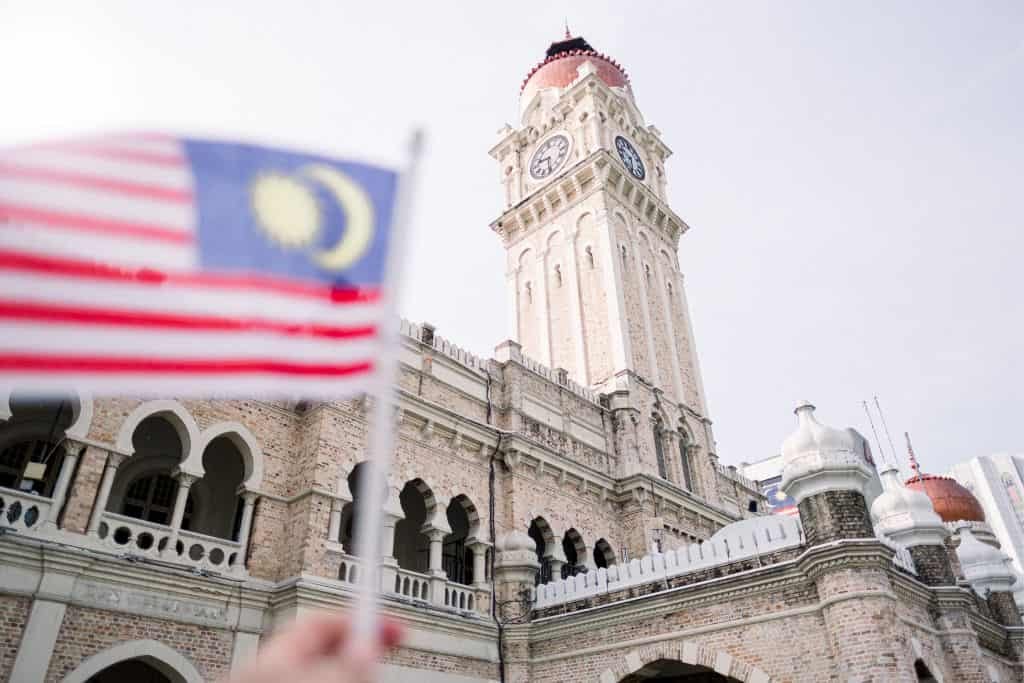 This image shows a person holding a Malaysian flag in front of a historic building with a tall clock tower and a red dome. The structure has ornate stonework and arched windows, standing out against a pale sky.
