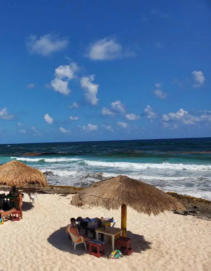 A serene beach scene with thatched-roof palapas providing shade over tables and chairs in the soft white sand. A small group of people sits under one of the palapas, enjoying the ocean view, while waves crash gently against the rocky shoreline. The deep blue sky, scattered with fluffy clouds, enhances the tropical and relaxing atmosphere on the east side of Cozumel.