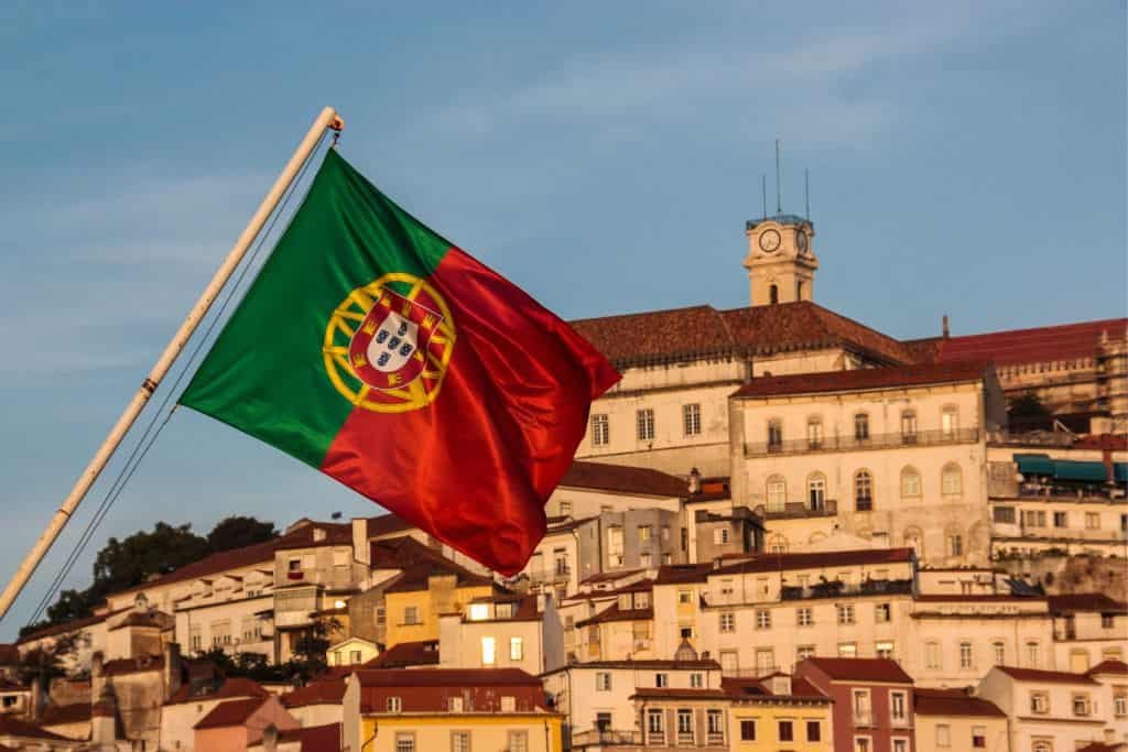 This image shows a Portuguese flag waving in the foreground with a hillside cityscape of historic buildings behind it. A clock tower rises above the rooftops, bathed in warm sunlight under a clear blue sky.
