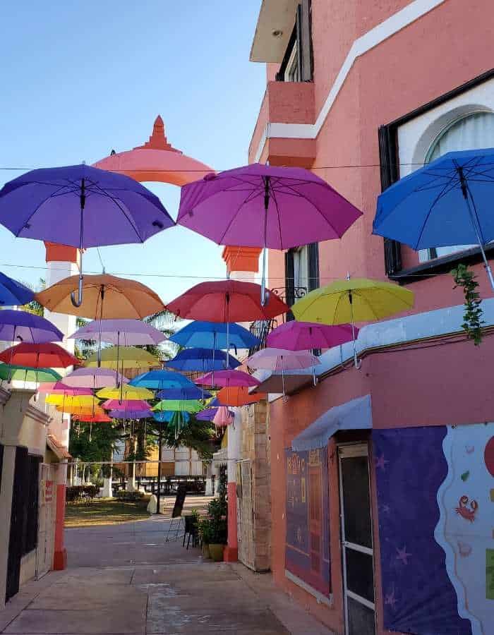 Narrow alley in Cozumel with a canopy of colorful umbrellas suspended overhead, creating a playful and vibrant atmosphere between pastel-colored buildings.