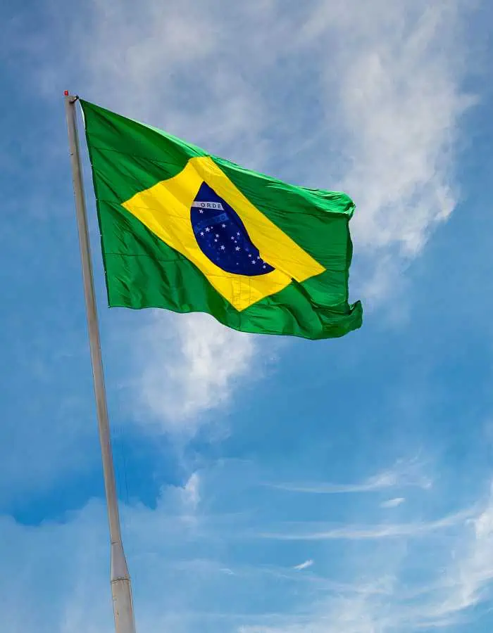 This image shows the Brazilian flag waving against a bright blue sky with wispy clouds. The green background, yellow diamond, and blue globe with stars are clearly visible on the flag.