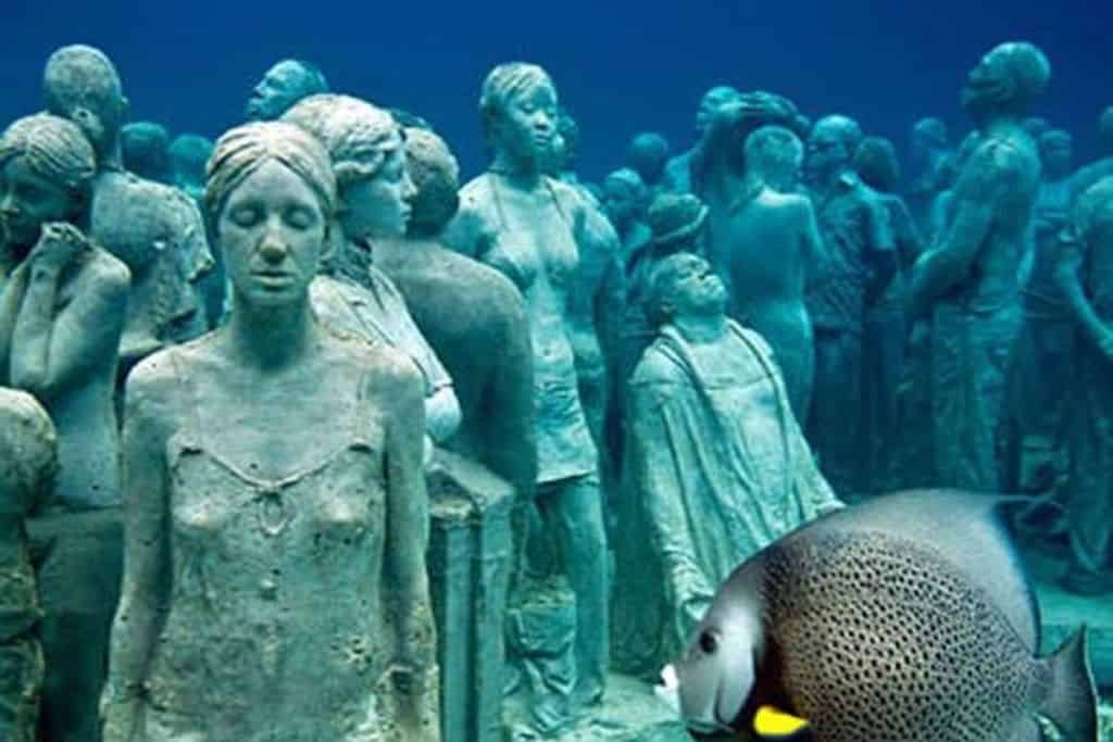 Underwater view of life-size human statues at the Cancún Underwater Museum (MUSA), surrounded by clear blue water with a fish swimming in the foreground.