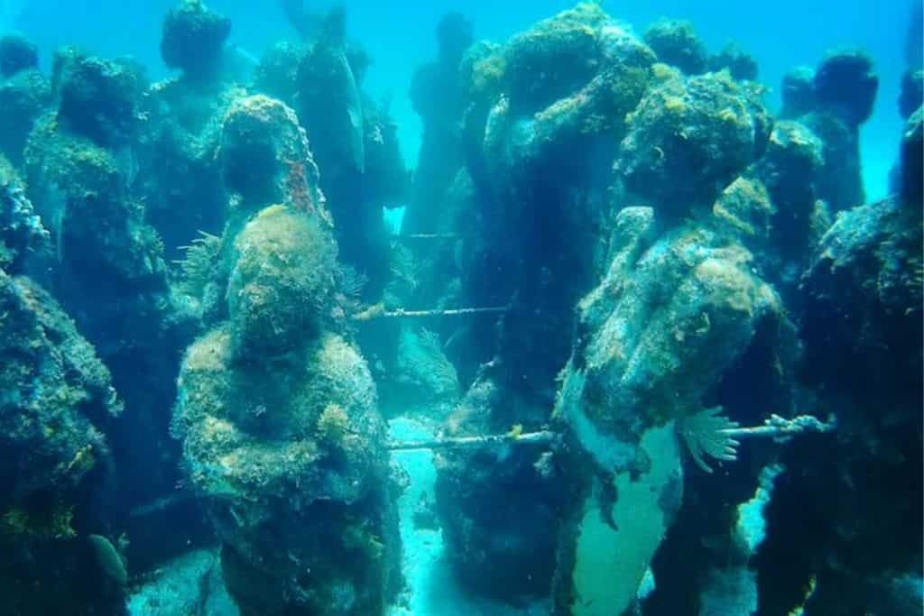 Underwater sculpture installation depicts human figures standing in a circle, covered in algae and marine growth. The statues create an eerie yet artistic scene on the ocean floor.