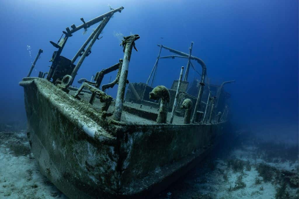 Sunken shipwreck rests on the ocean floor, covered in algae and marine growth. The skeletal frame tilts slightly in the clear blue water, creating an eerie underwater scene.