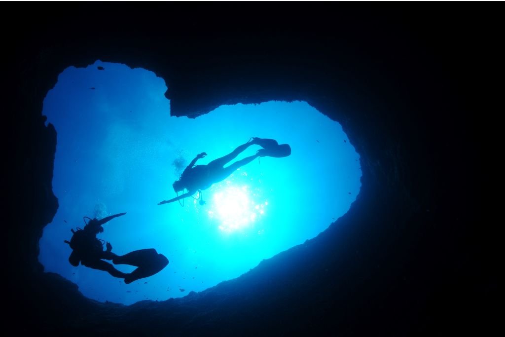 Two divers are silhouetted against bright blue water as they swim through an underwater cave opening. Sunlight filters through the entrance, creating a striking contrast with the dark cave interior.