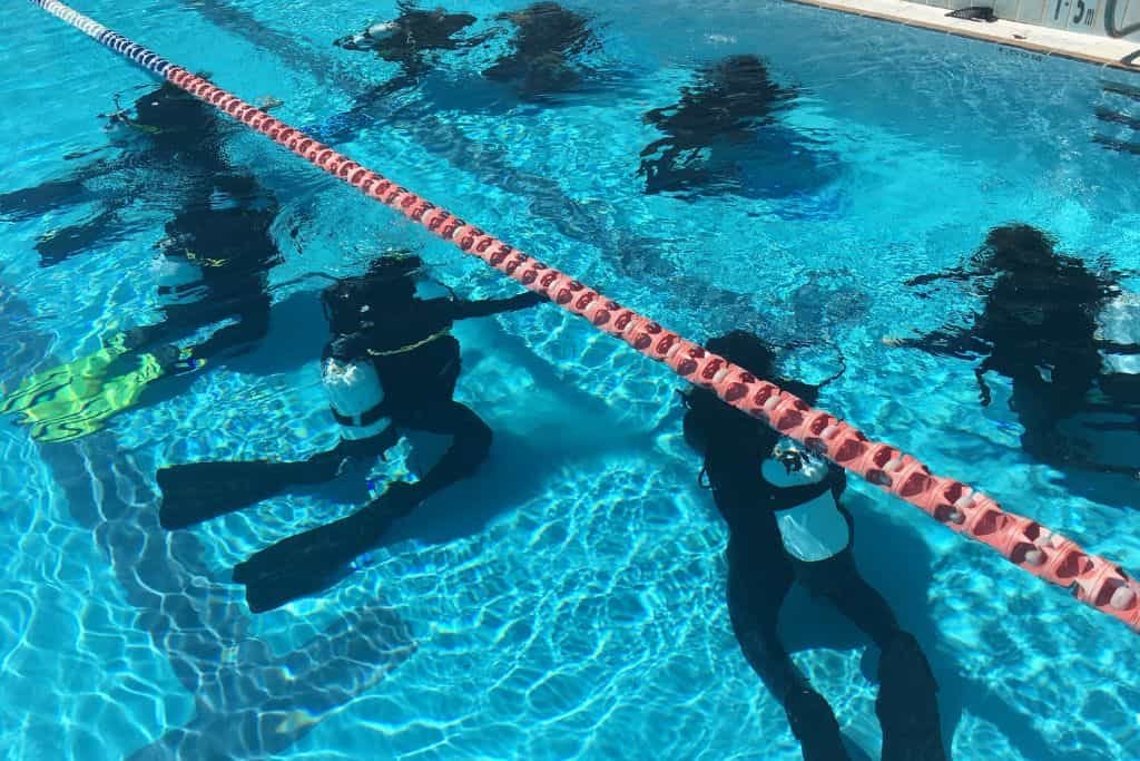 Group of scuba divers practice underwater skills in a swimming pool, kneeling along a lane divider. Clear blue water reveals their tanks, fins, and wetsuits during training.