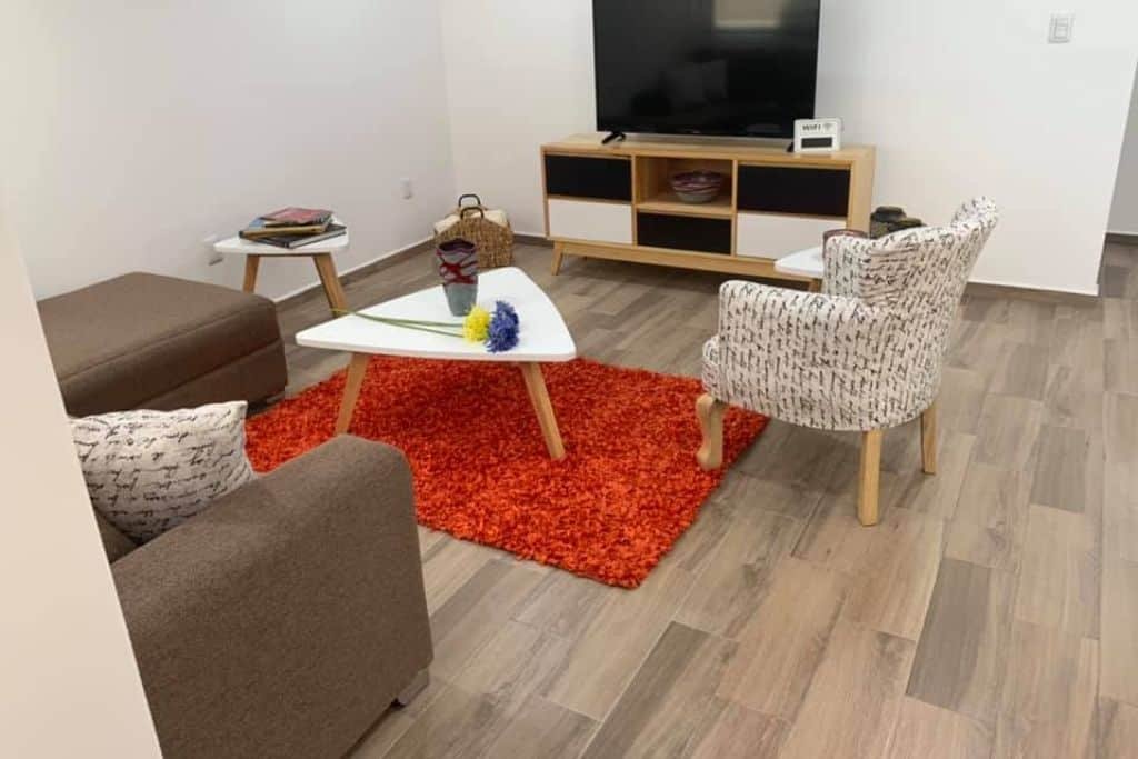 A cozy living room features a brown sofa, patterned armchair, and white triangular coffee table on a bright orange rug. A wooden TV stand with storage compartments holds a large flat-screen television against a plain white wall.