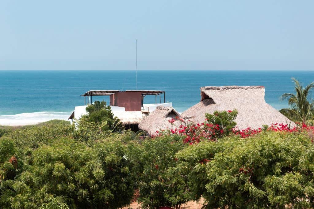 Thatched-roof buildings peek out from dense green foliage overlooking a calm blue ocean. The scene captures a tropical coastal setting with bright, clear skies.