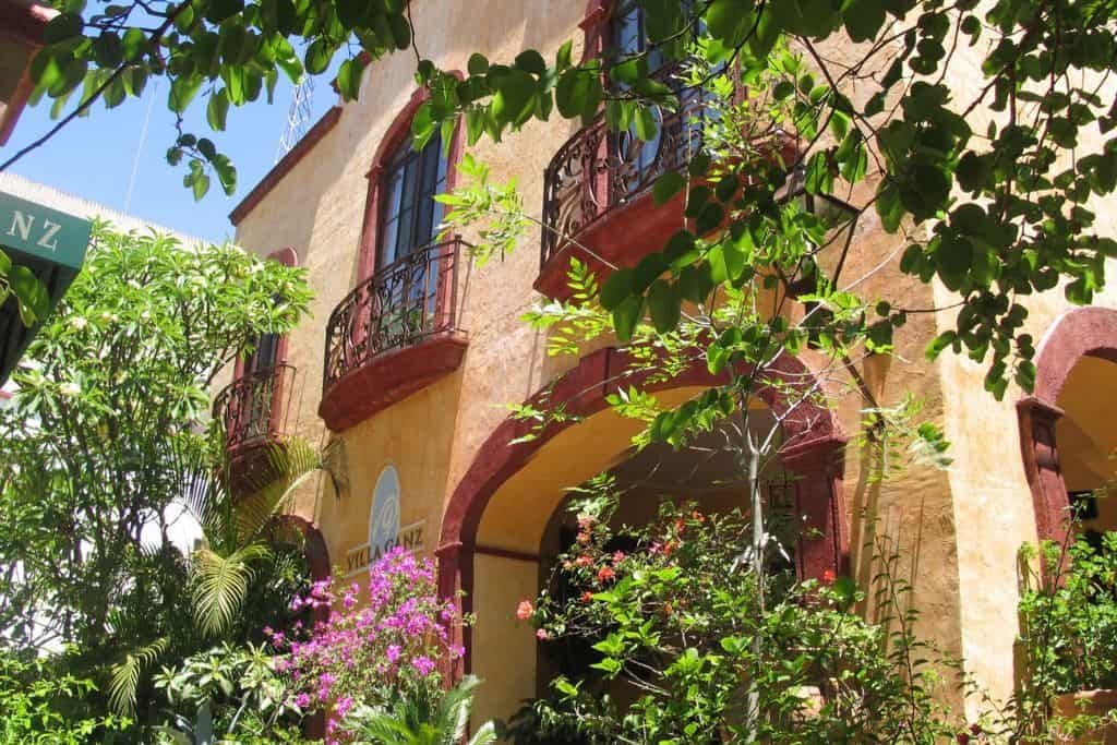 A two-story building with warm yellow and red tones features arched doorways and wrought-iron balconies. Lush greenery and pink flowers surround the exterior, creating a vibrant, shaded courtyard feel.