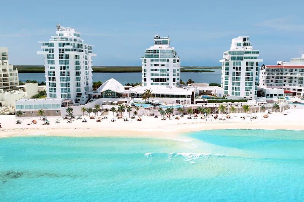 A row of modern beachfront hotels with white facades faces a wide stretch of white sand and turquoise water. Lounge chairs and umbrellas are set up along the shoreline under a clear sky.